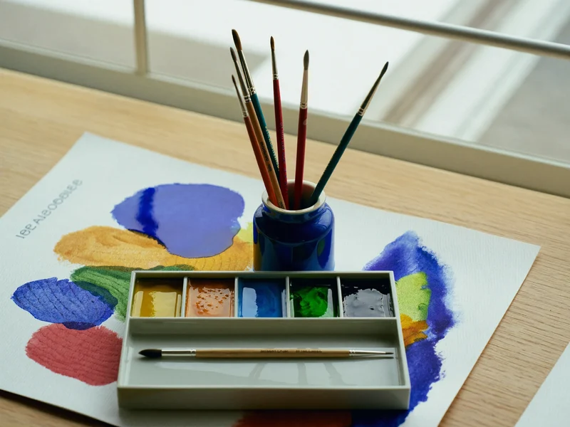 Illustrative watercolour paints, brushes in a jar and a loose wash on textured paper